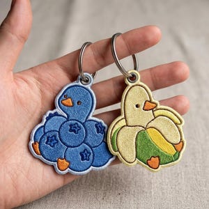 May include: Two embroidered keychains featuring stylized ducks. One duck is blue and shaped like a cluster of blueberries with an orange beak and feet. The other duck is yellow and shaped like a peeled banana with a green peel, orange beak, and feet.