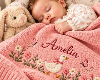 May include: A soft pink knitted baby blanket featuring embroidered details of a white goose with a pink bow, daisies, and butterflies. The name Amelia is stitched in dark pink script. A plush beige bunny toy sits beside the blanket.