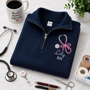 May include: A navy blue quarter-zip sweatshirt featuring an embroidered pink stethoscope shaped like a bow, with the name Lily and the title RN stitched below in white. The garment has a metal zipper and a Gildan brand label at the inner neckline.