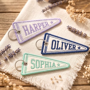 May include: Three triangular embroidered keychains with metal rings. The top keychain is lavender with the name HARPER, the middle is navy blue with the name OLIVER, and the bottom is mint green with the name SOPHIA. Each features a star icon.