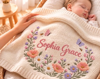 May include: A cream-colored knitted baby blanket featuring an embroidered floral wreath with pink, white, and purple flowers, small bees, and butterflies. The name Sophia Grace is embroidered in pink script in the center of the wreath.