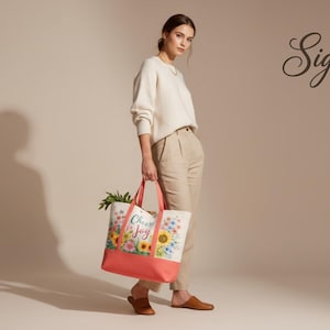 May include: A woman holds a tote bag with a floral design and the words "Choose Joy". The bag has a coral-coloured base and handles. The background is a neutral beige.