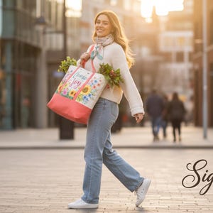 May include: A person walks along a city street carrying a tote bag. The bag is white with a floral design and the words "Choose Joy". The bag has a coral-coloured bottom. They wear a white jumper, jeans, and white trainers.