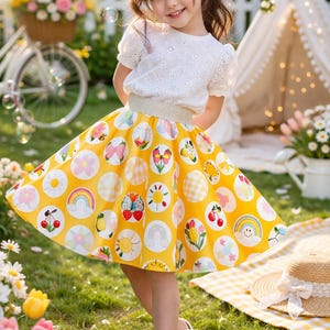 May include: A young girl wearing a white eyelet top and a yellow skirt adorned with colorful circular patterns. The skirt features rainbows, cherries, and other designs. She is wearing white sandals and standing on grass.