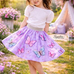 May include: A young girl wearing a white eyelet top and a lavender skirt adorned with colorful butterfly illustrations. The skirt has a silver waistband. She is wearing white sandals. The image is taken outdoors in a garden setting.