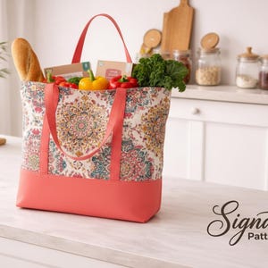 May include: A coral and patterned tote bag filled with groceries, including a baguette, vegetables, and packaged goods. The bag has coral handles and a coral bottom. The bag is sitting on a white countertop.