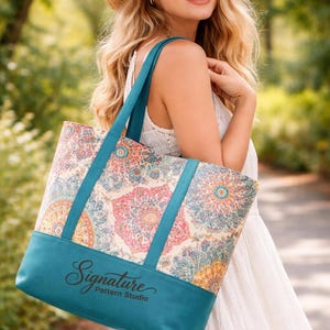 May include: A teal tote bag with a colourful floral pattern and teal straps. The bag has the text "Signature Pattern Studio" on the front. The person in the image is wearing a white dress and a straw hat.