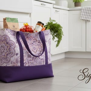 May include: A large tote bag with a purple and white paisley pattern, filled with groceries. The bag has a solid purple base and matching handles. The bag contains a bag of flour, tomatoes, carrots, and a jar of sauce.