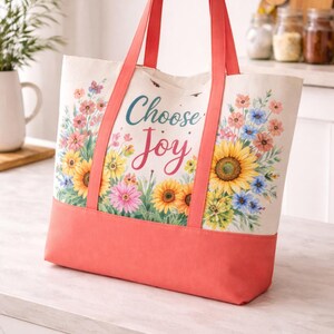 May include: A canvas tote bag with coral handles and a coral base. The bag displays a floral design with sunflowers and the words "Choose Joy". The Signature Pattern Studio logo is in the upper left corner.