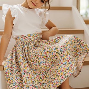 May include: A young girl wearing a white top with ruffled sleeves and a floral print skirt. The skirt features a pattern of small flowers in various colors. The girl is sitting on wooden stairs.