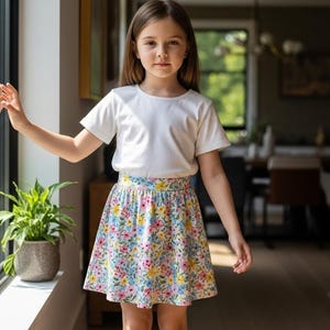 May include: A young person wearing a white t-shirt and a floral print skirt. The skirt features a pattern of colorful flowers on a light background. The outfit is suitable for casual wear. The image is well-lit, with natural light.