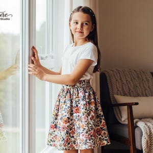 May include: A young person wearing a white t-shirt and a floral print skirt stands near a window. The skirt features a pattern of red, pink, and yellow flowers. The text "Signature Pattern Studio" is visible in the upper left corner.