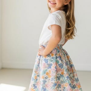 May include: A young girl wearing a white t-shirt and a floral skirt. The skirt features a pattern of blue, pink, and yellow flowers on a white background. A floral headband complements the outfit. The text "Signature Pattern Studio" is in the upper left corner.
