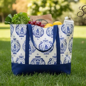 May include: A tote bag filled with groceries rests on grass. The bag features a cream background with a blue vase pattern, a solid blue base and straps. It contains lettuce, grapes, lemons, and other items. The bag is outdoors.