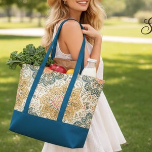 May include: A patterned tote bag with a teal base and straps, filled with groceries. The bag's upper section features a gold, teal, and cream design. The bag is being carried outdoors, with kale, apples, and a bottle of milk visible.