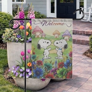 Może przedstawiać: Flaga ogrodowa z Snoopy'm i Woodstockiem, z napisem "Welcome". Flaga ma kolorowy wzór z domkami z grzybów, kwiatami i zielonym tłem. Flaga jest wyświetlana na czarnym metalowym stojaku.