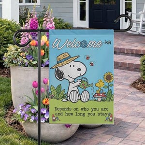 Może przedstawiać: Niebieska flaga ogrodowa z Snoopy podlewającym roślinę, z Woodstockiem w pobliżu. Flaga głosi "Welcome-ish" i "Zależy od tego, kim jesteś i jak długo zostajesz". Flaga jest na czarnym metalowym stojaku.