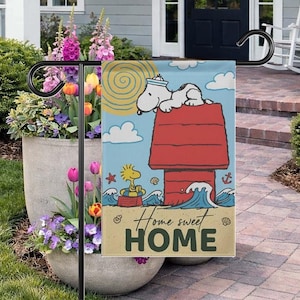 May include: A garden flag with Snoopy in a sailor hat on his red doghouse, and Woodstock in a boat. The flag has a blue sky background with clouds and waves. The text reads "Home sweet HOME".