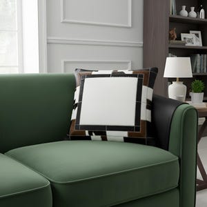 May include: A decorative square pillow with a geometric pattern. The pillow features a white center square framed by a black border, a brown border, and a black and white cowhide-like pattern. The pillow is placed on a green sofa.