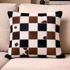 May include: Square cushion with a patchwork design. The cushion features a checkerboard pattern of black, white, and brown squares. The cushion has a black border and is made of a soft, textured material.