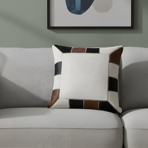May include: Square decorative pillow with a white center and a patchwork border of black, brown, and white sections. The pillow is placed on a light gray sofa, with a framed abstract artwork in the background.