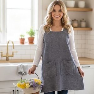 Peut inclure: Une femme porte un tablier gris avec deux grandes poches sur une chemise blanche à manches longues. Elle tient un panier de fleurs. La cuisine comprend un évier blanc, un robinet en laiton et des étagères en bois.