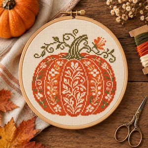 Peut inclure: Un tambour à broder circulaire en bois contenant un motif au point de croix représentant une grande citrouille orange avec des détails floraux blancs et des vignes vertes. Le motif est centré sur un tissu crème avec une tige vert foncé.