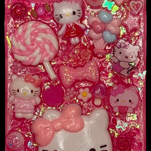 Puede incluir: Funda de tel&eacute;fono rosa decorada con varios adornos 3D. Presenta m&uacute;ltiples figuras de Hello Kitty, lazos, corazones, flores y una piruleta. La funda est&aacute; llena de purpurina rosa y otros elementos decorativos.