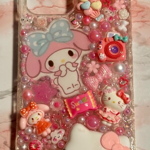 Puede incluir: Funda de tel&eacute;fono transparente decorada con personajes de dibujos animados rosas y blancos, caramelos y cuentas. Presenta a Hello Kitty, My Melody y una c&aacute;mara rosa. La palabra "Sweet" es visible en un envoltorio de caramelos.