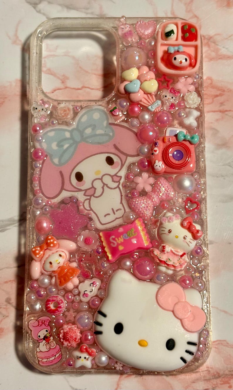 Puede incluir: Funda de tel&eacute;fono transparente decorada con personajes de dibujos animados rosas y blancos, caramelos y cuentas. Presenta a Hello Kitty, My Melody y una c&aacute;mara rosa. La palabra "Sweet" es visible en un envoltorio de caramelos.