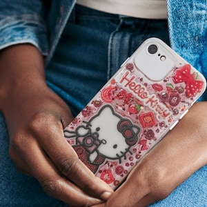 Puede incluir: Una funda transparente para tel&eacute;fono con un dise&ntilde;o de Hello Kitty. La funda tiene un borde blanco y un fondo rosa con detalles en rojo, incluyendo un lazo, cerezas y las palabras "Hello Kitty".