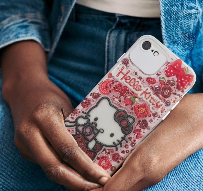 Puede incluir: Una funda transparente para tel&eacute;fono con un dise&ntilde;o de Hello Kitty. La funda tiene un borde blanco y un fondo rosa con detalles en rojo, incluyendo un lazo, cerezas y las palabras "Hello Kitty".