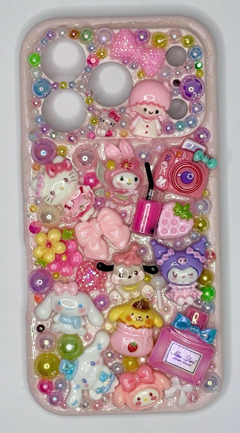 Puede incluir: Funda de tel&eacute;fono rosa decorada con varios abalorios 3D, incluyendo Hello Kitty, My Melody y otros personajes de dibujos animados. La funda est&aacute; adornada con cuentas, lazos y un frasco de perfume, creando un dise&ntilde;o lindo y colorido.