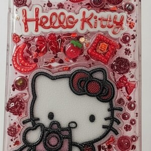 Puede incluir: Funda de tel&eacute;fono transparente con un dise&ntilde;o rosa y rojo que presenta a Hello Kitty sosteniendo una c&aacute;mara. La funda est&aacute; decorada con lazos rojos, fresas y purpurina. Las palabras "Hello Kitty" son rojas.
