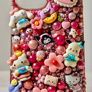 Puede incluir: Una funda de tel&eacute;fono rosa adornada con varios dijes de personajes de dibujos animados y cuentas rosas. La funda presenta a Hello Kitty, Cinnamoroll y otros personajes. La funda tiene un acabado brillante y texturizado.