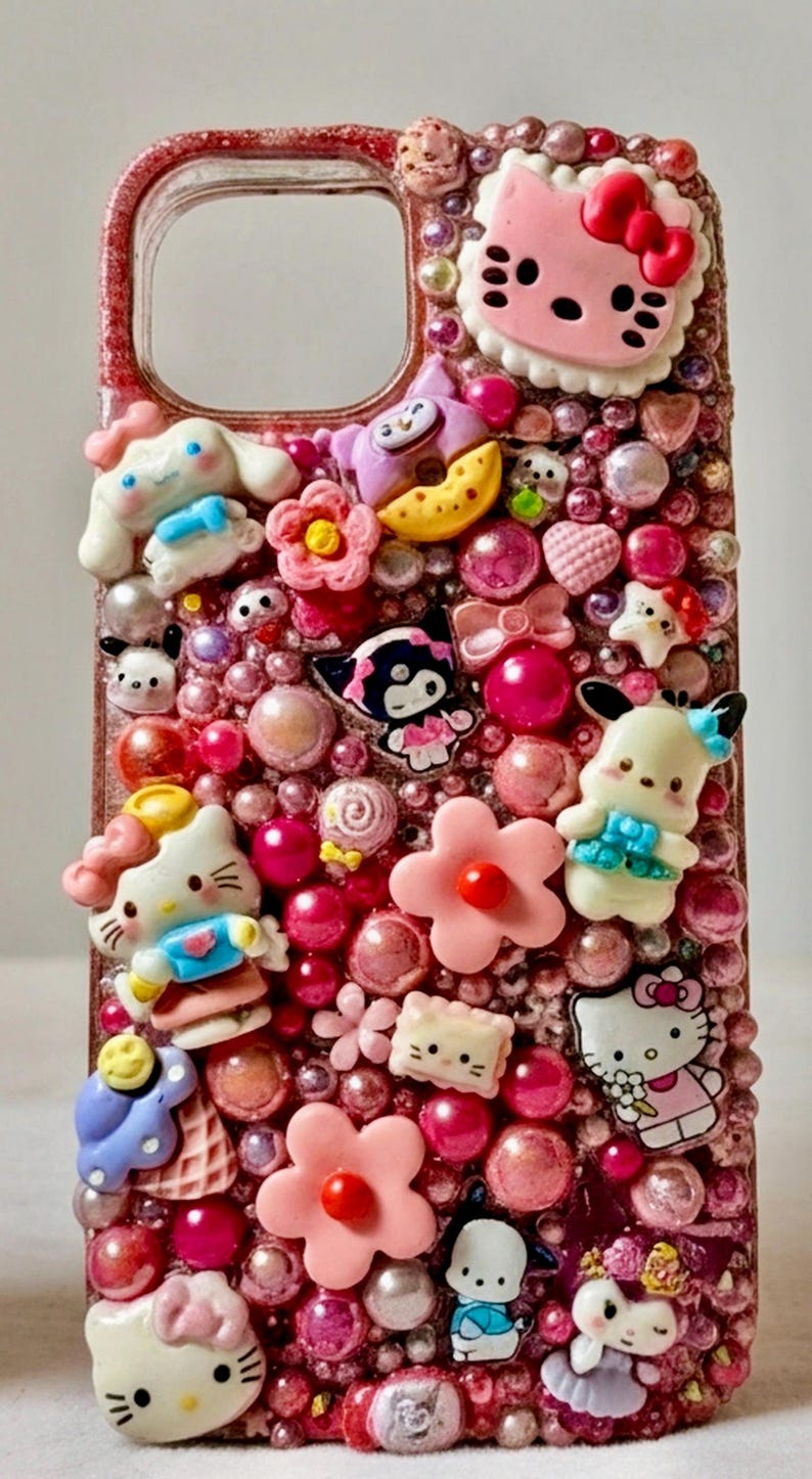 Puede incluir: Una funda de tel&eacute;fono rosa adornada con varios dijes de personajes de dibujos animados y cuentas rosas. La funda presenta a Hello Kitty, Cinnamoroll y otros personajes. La funda tiene un acabado brillante y texturizado.