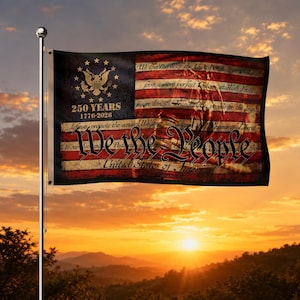 Könnte beinhalten: Eine rechteckige Flagge mit einem dunkelblauen Kanton, einem goldenen Adleremblem, dem Text 250 YEARS 1776-2026 sowie roten und weißen Streifen. Die Flagge zeigt den Text We the People und Auszüge aus der US-Verfassung in schwarzer Schrift.