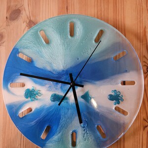 Op de afbeelding: Ronde wandklok met een blauw en wit oceaanontwerp. De wijzerplaat heeft een wervelend patroon met ingebedde zeedieren, waaronder kwallen, een octopus en een pijlstaartrog. Zwarte wijzers en uurmarkeringen maken het ontwerp compleet.