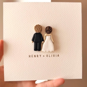 May include: A square white card featuring two small, three-dimensional figures standing side-by-side. One figure has light brown hair and wears a black suit, while the other has dark brown hair and a white dress. The text HENRY + OLIVIA is printed below.