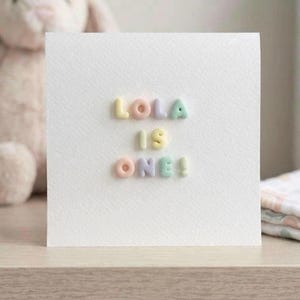 May include: A square white greeting card with textured paper featuring the raised, pastel-coloured letters spelling out "LOLA IS ONE!" in three rows. The letters are in shades of yellow, pink, purple, and green, arranged in a centred block.