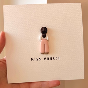 May include: A square greeting card featuring a three-dimensional clay figure of a person with dark brown hair tied in a low ponytail, wearing a white long-sleeved top and light pink wide-leg trousers. The text MISS MUNROE is printed in black.