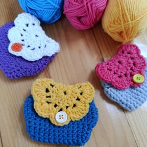 Könnte beinhalten: Nahaufnahme von mehreren handgefertigten gehäkelten Münzbörsen in verschiedenen Farben, darunter Blau, Lila und Gelb. Jede Börse hat eine kontrastierende gehäkelte Klappe und einen Knopfverschluss. Bunte Wollknäuel sind im Hintergrund zu sehen.