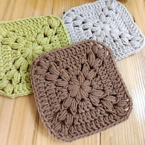 Puede incluir: Tres posavasos hechos a mano de ganchillo en tonos verdes, grises y marrones. Cada posavasos cuadrado presenta un diseño floral en el centro. Los posavasos están hechos de un hilo suave y están colocados sobre una superficie de madera.