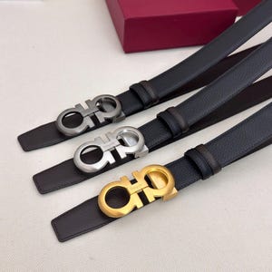 May include: Three black leather belts with silver and gold-tone buckles. The buckles feature a double-Gancio design. A red box with the brand name is visible in the background.