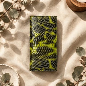 Può includere: Un portafoglio rettangolare caratterizzato da un motivo a pelle di serpente lucido e testurizzato in verde lime vibrante e nero profondo. La superficie presenta un aspetto distintivo a scaglie con una finitura lucida e contrastante.