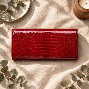 May include: A rectangular wallet with a glossy, deep red finish and a textured crocodile skin pattern. The wallet features a clean, structured design with a smooth, reflective surface that highlights the intricate embossed scale details.