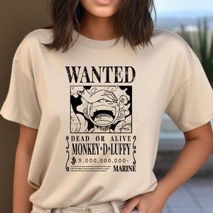 May include: Beige t-shirt featuring a black and white graphic of a "Wanted" poster. The poster includes the text "WANTED", "DEAD OR ALIVE", and "MONKEY D. LUFFY". The graphic also includes a drawing of a character with his hand over his eyes.