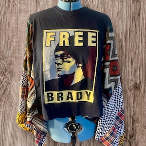 May include: A black long-sleeved shirt with a graphic print of a man's face with the text "FREE BRADY" on the front. The sleeves are made of a patchwork of colorful fabrics.
