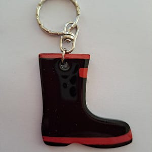 Op de afbeelding: Een zwarte en rode laars sleutelhanger. De sleutelhanger heeft een glanzende zwarte laars met rode accenten op de bovenkant en zool. De laarsvormige bedel is bevestigd aan een zilverkleurige sleutelring.