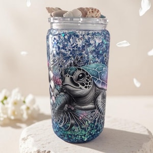 May include: A decorative jar with a sea turtle design. The jar features a detailed illustration of a sea turtle surrounded by aquatic plants and glitter. The lid is adorned with seashells. The jar is placed on a white stone surface.